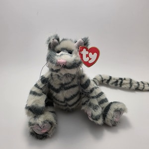 Op de afbeelding: Een pluche kat met grijze en witte strepen, een roze neus en voetzolen, en een rood hartvormig label. De kat heeft een lange staart en zit.