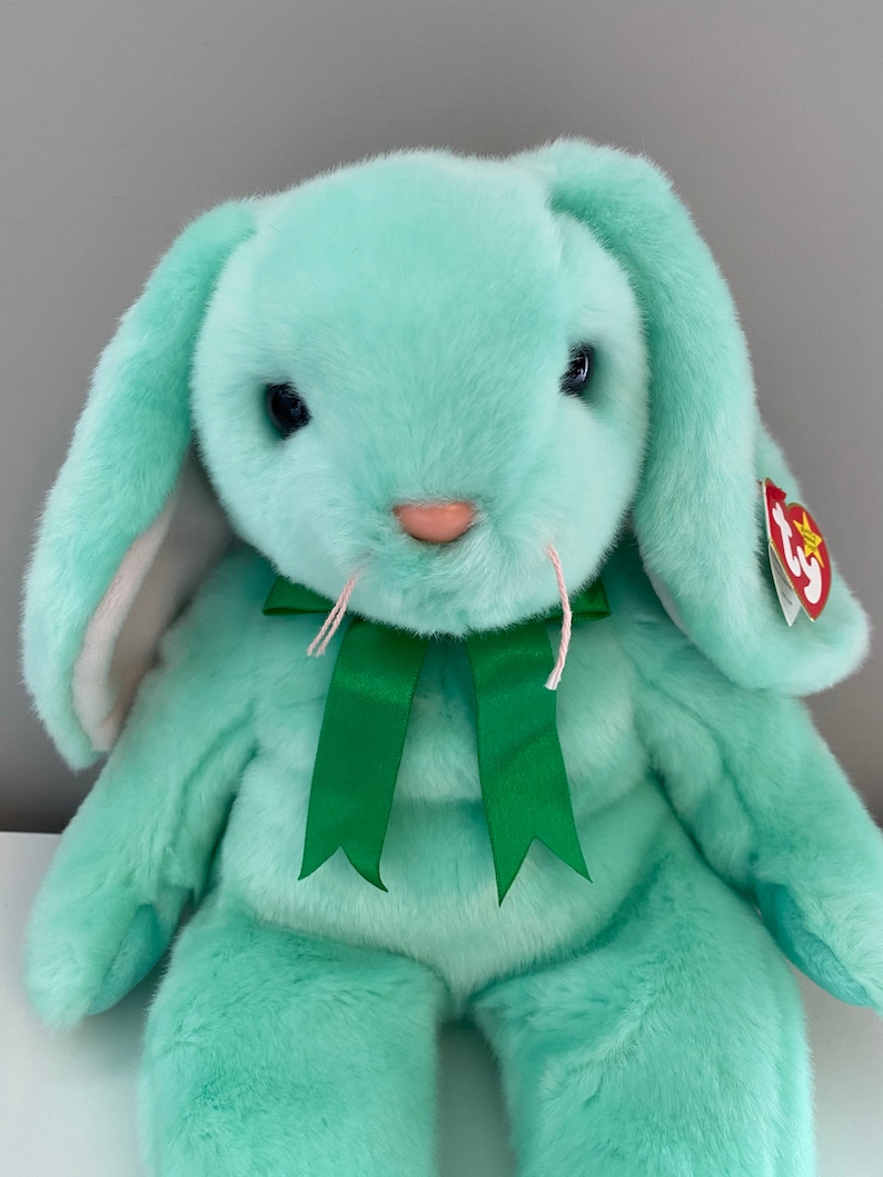 Puede incluir: Un conejito de peluche verde menta con largas orejas ca&iacute;das y un lazo de cinta verde. El conejito tiene ojos negros, bigotes rosas y una nariz rosa. Una etiqueta roja est&aacute; sujeta a la oreja.