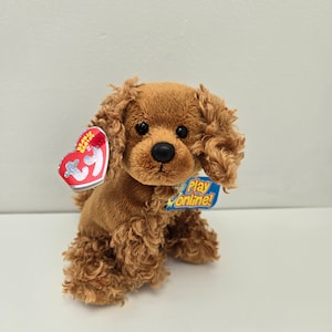May include: A brown plush toy dog with curly fur. The dog is sitting with its head tilted to the side. The dog has a red tag with the words "Beanie Babies" and a blue tag with the words "Play online!"