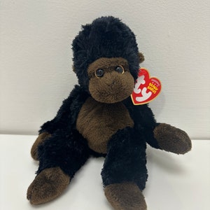 May include: A plush gorilla toy with black fur and brown accents. The toy has a brown face and feet, with a red heart-shaped tag attached. The gorilla is sitting with its arms and legs spread out.