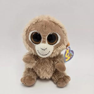 May include: A brown and white plush monkey with large, black eyes and a Ty tag. The monkey is sitting with its legs crossed.
