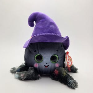 May include: A plush toy octopus with large green eyes, rosy cheeks, and a smiling mouth, wearing a purple witch hat. The octopus has gray and multi-colored tentacles. A red and white tag is attached.