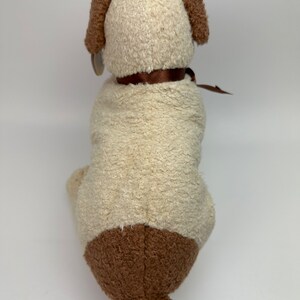 Op de afbeelding: Een pluche honden speelgoed met een cr&egrave;mekleurig lichaam en bruine oren, poten en staart. Het speelgoed draagt een bruin lint om zijn nek.