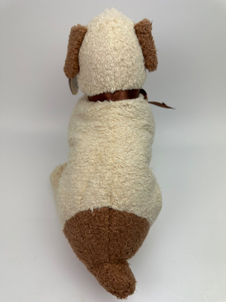 Op de afbeelding: Een pluche honden speelgoed met een cr&egrave;mekleurig lichaam en bruine oren, poten en staart. Het speelgoed draagt een bruin lint om zijn nek.