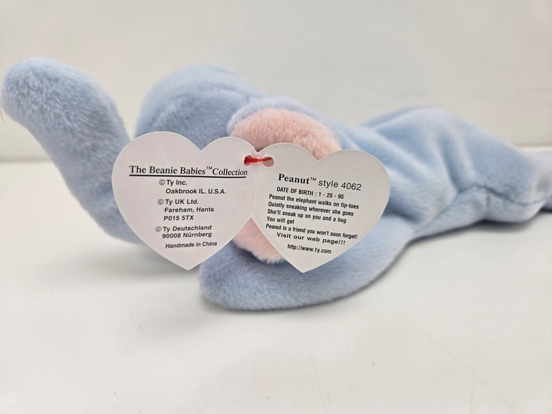 May include: A light blue Beanie Baby elephant named Peanut with a pink nose and a red heart-shaped tag. The tag includes the name Peanut, the style number 4062, and the date of birth 1-25-95. The tag also includes information about the manufacturer, Ty Inc., and the country of origin, China.