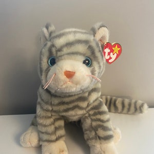 以下が含まれることがあります： 青い目とピンクの鼻を持つ、グレーと白のストライプのぬいぐるみ猫。ぬいぐるみには、耳に赤いハート型のタグが付いています。猫は白い面に座っています。