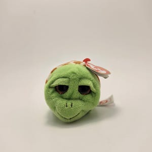 May include: A green plush turtle toy with a brown and tan shell pattern. The turtle has large, glittery brown eyes and a small red tag attached to its head. The toy is made of soft, fuzzy material.