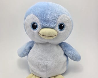 Può includere: Un giocattolo di peluche di pinguino blu chiaro con pancia bianca e piedi gialli. Il pinguino ha occhi neri e un becco giallo.