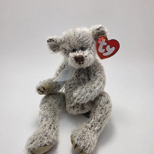 Può includere: Un orsacchiotto di peluche con pelliccia arruffata grigia e bianca maculata, caratterizzato da un naso cucito marrone scuro e cuscinetti delle zampe in tessuto color cuoio. L'orso indossa un nastro azzurro trasparente al collo e un'etichetta a cuore.