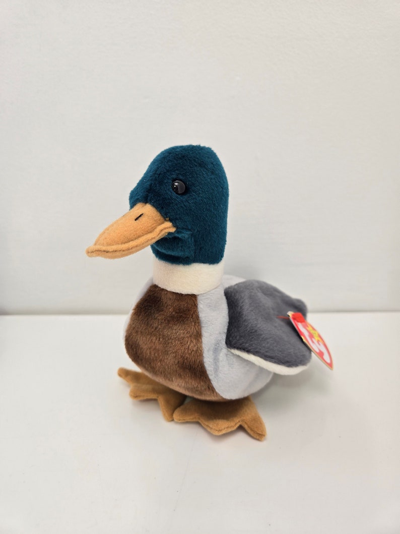 Pu&ograve; includere: Un'anatra di peluche blu-verde e marrone con una striscia bianca intorno al collo. L'anatra ha un becco marrone e occhi neri. L'anatra &egrave; seduta su una superficie bianca. L'anatra ha un'etichetta Ty rossa e bianca attaccata alla sua ala.