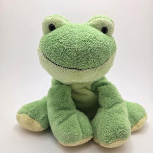 May include: A plush green frog toy with a light green body and a cream-colored belly and feet. The frog has black eyes and a stitched black smile. The soft toy is sitting on a white surface.