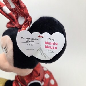 May include: A Minnie Mouse soft toy from the Beanie Buddies Collection. The toy features a red dress with white polka dots, a black head, and a red bow with white polka dots. The Disney tag is visible.
