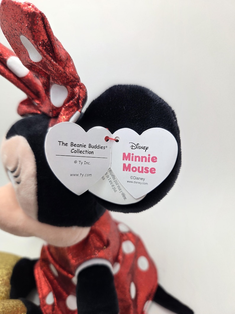 May include: A Minnie Mouse soft toy from the Beanie Buddies Collection. The toy features a red dress with white polka dots, a black head, and a red bow with white polka dots. The Disney tag is visible.