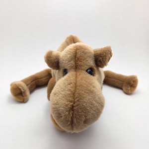 May include: A plush camel toy in shades of brown, lying on a white surface. The toy has a soft, textured appearance with detailed features, including small ears, dark eyes, and outstretched legs.