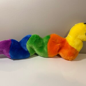 Op de afbeelding: Een pluche regenboogworm speelgoed met een geel hoofd en zwarte ogen. De worm is gemaakt van zacht, kleurrijk materiaal en is perfect om te knuffelen.