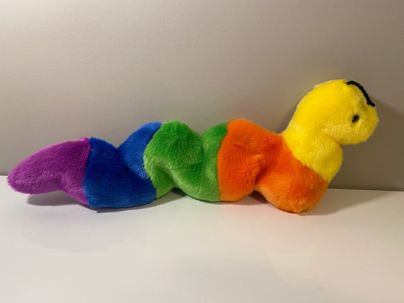 Op de afbeelding: Een pluche regenboogworm speelgoed met een geel hoofd en zwarte ogen. De worm is gemaakt van zacht, kleurrijk materiaal en is perfect om te knuffelen.