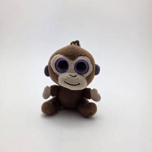 May include: A small, plush monkey toy. It has brown fur, large, round eyes with purple accents, and small ears. The monkey is sitting upright with its arms and legs extended. The toy has a friendly, cartoonish appearance.