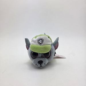 May include: A soft toy dog in grey, with large dark eyes and a green and white cap. The cap has a Paw Patrol logo. The toy has grey ears and a black nose. A small tag is attached.
