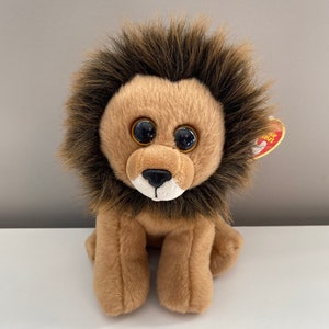 May include: A plush lion toy with a tan body, brown mane, and large, round, black-pupiled eyes. The lion is sitting upright with its paws forward. A red tag is attached to its ear.