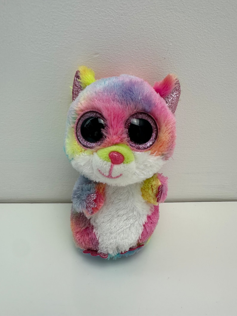 May include: A colorful plush toy with large, glittery eyes. The toy is pink, blue, yellow, and green with a white belly. It has a cute, cartoonish face.