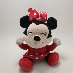 May include: A plush Minnie Mouse doll with a red dress and bow, both adorned with white polka dots. The doll has black ears, a black nose, and a smiling face. The doll's arms and legs are red.