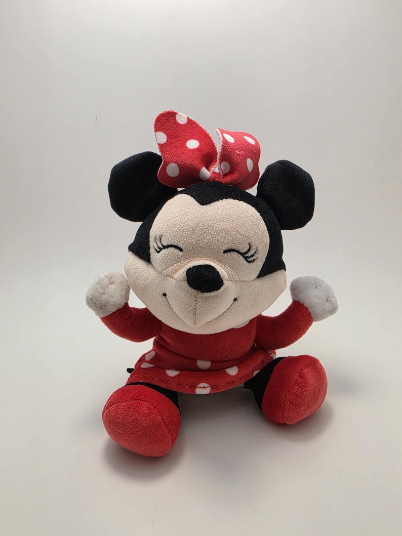 May include: A plush Minnie Mouse doll with a red dress and bow, both adorned with white polka dots. The doll has black ears, a black nose, and a smiling face. The doll's arms and legs are red.