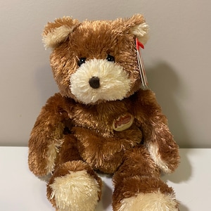 May include: A brown and cream plush teddy bear with a white face and a red tag. The bear is sitting on a white surface.