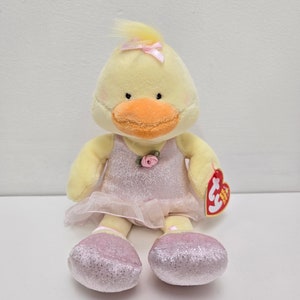 May include: A plush yellow duck wearing a pink tutu and ballet slippers. The duck has an orange beak, a small pink bow on its head, and a pink rose on its dress. A red heart-shaped tag is attached.