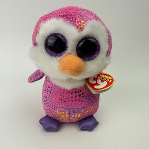 May include: Pink and white penguin plush toy with sparkly pink and gold fabric. The toy has large, black eyes with purple eyelids and a small orange beak. The toy has a purple tag that says "Ty Beanie Boos".