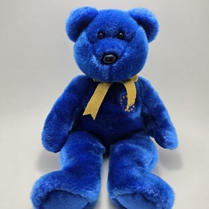 May include: A plush teddy bear with bright blue fur, black plastic eyes, and a black nose. A gold ribbon is tied around its neck. On the chest, there is an embroidered circular emblem featuring a ring of yellow stars on a blue background.