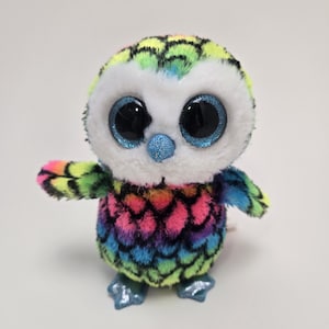 May include: A colorful plush owl toy with large, glittery blue eyes and a blue glittery beak. The owl's body is covered in a rainbow-colored scale pattern, and it has small wings and feet.