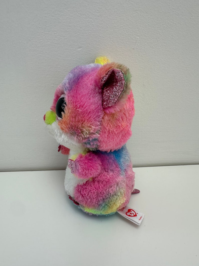May include: A colorful tie-dye plush toy hamster with large black eyes and a pink glittery ear. The toy has a white belly and a red heart tag that says "Ty".
