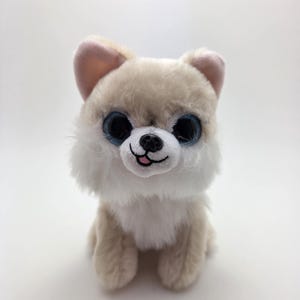 May include: A soft toy dog with tan and white fur, large blue eyes, and pink inner ears. The stuffed animal is sitting upright, with its paws visible. The dog has a black nose and a small, smiling mouth.