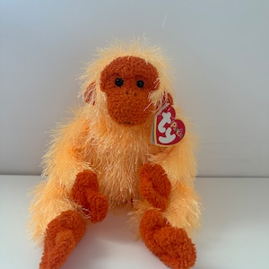 May include: A soft orangutan toy in shades of orange and brown, with a heart-shaped tag. The toy is depicted sitting with its legs extended. The orangutan is made from a soft, fuzzy material.