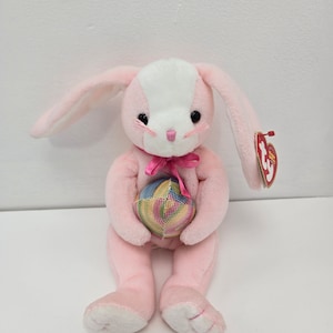 May include: Pink plush bunny holding a colorful Easter egg. The bunny has white fur on its face and a pink bow. The bunny is sitting on a white surface.