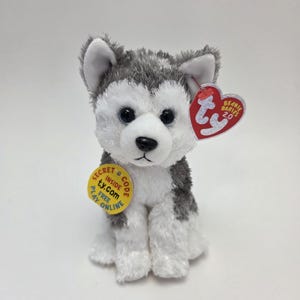 Op de afbeelding: Een grijze en witte pluche husky-hond die zit met een rood en wit Ty Beanie Babies 2.0-label en een geel label met de tekst "Geheime code binnen ty.com Gratis online spelen".
