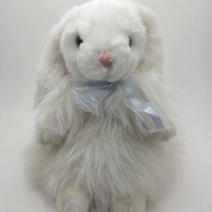 May include: A white plush rabbit with long ears and a light blue ribbon bow. The rabbit has a pink nose, blue eyes, and whiskers. The soft, fluffy fur gives it a cuddly appearance. A charming soft toy.