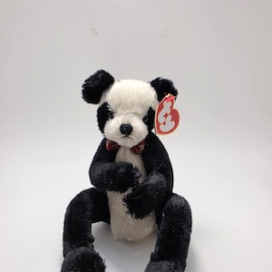 Può includere: Un orsetto panda di peluche con pelliccia bianca e nera, caratterizzato da un piccolo papillon rosso al collo e un'etichetta Ty rossa a forma di cuore attaccata all'orecchio sinistro. L'orso è seduto con arti neri e busto bianco.