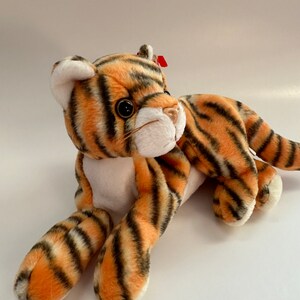 Op de afbeelding: Een pluche tijger speelgoed met oranje en zwarte strepen. Het speelgoed heeft een witte buik en een rood label op zijn oor.