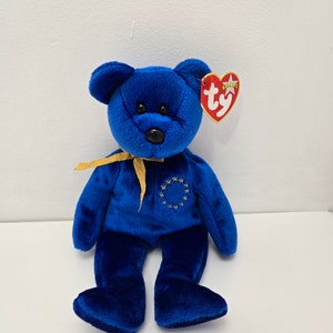 May include: Blue Beanie Baby bear with a gold ribbon and a gold embroidered circle of stars on its chest. The bear has a red Ty heart tag attached to its ear.