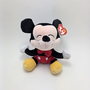 May include: A plush Mickey Mouse toy with a red bow tie, red shorts with white buttons, yellow shoes, and black ears. The toy has a smiling face and white gloves. A red tag is attached to the ear.