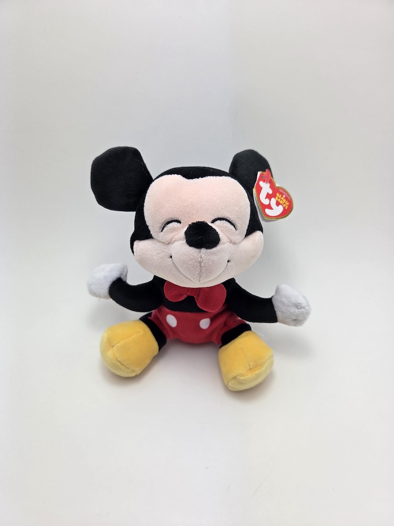 May include: A plush Mickey Mouse toy with a red bow tie, red shorts with white buttons, yellow shoes, and black ears. The toy has a smiling face and white gloves. A red tag is attached to the ear.