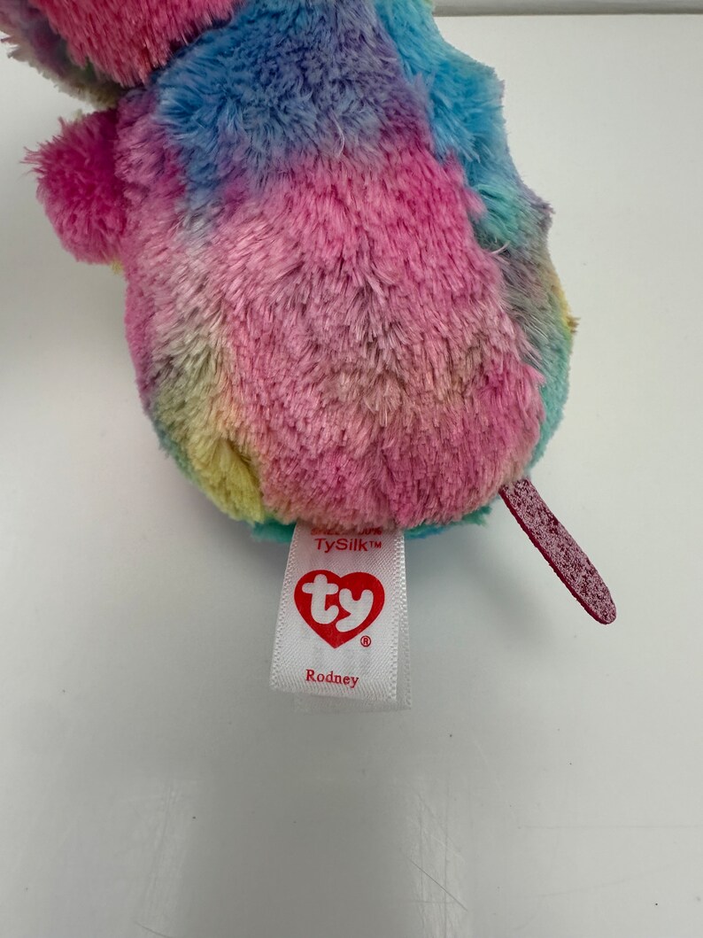 May include: A rainbow tie-dye plush toy with a red tail. The toy has a TySilk tag with the Ty logo and the name Rodney.