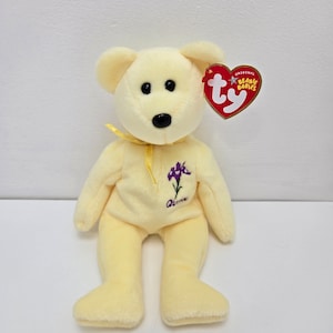 May include: A yellow plush beanie baby bear with a purple flower embroidered on its chest. The bear is sitting with its legs crossed and has a red Ty tag attached to its ear. The tag says "Original Beanie Babies".