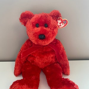Puede incluir: Un oso de peluche rojo con ojos negros y nariz negra. El oso tiene una cinta roja alrededor del cuello y una etiqueta en forma de corazón en la oreja. El oso está sobre una superficie blanca.