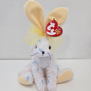 May include: A white and blue speckled beanie baby bunny with yellow ears and a yellow bow. The bunny is sitting with its paws tucked under its body. The bunny has a red Ty Beanie Babies tag attached to its ear.