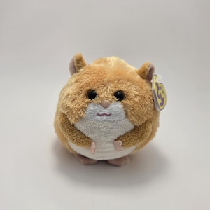 May include: A soft hamster toy in shades of brown and cream. It has a rounded shape, large black eyes, and small ears. The toy has a pink nose and paws. A small tag is attached to the ear.
