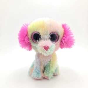 Può includere: Un colorato peluche a forma di cane con grandi occhi glitterati. Il cane ha una pelliccia color arcobaleno con accenti rosa sulle orecchie e un naso rosa. Il giocattolo è seduto in posizione eretta, mostrando la sua morbida consistenza.