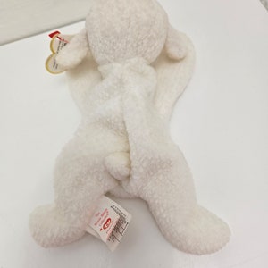 May include: A white plush lamb toy from The Beanie Babies Collection. The lamb has a soft, textured exterior, floppy ears, and a small tail. A tag is attached to the lamb's ear. The toy is lying on a white surface.