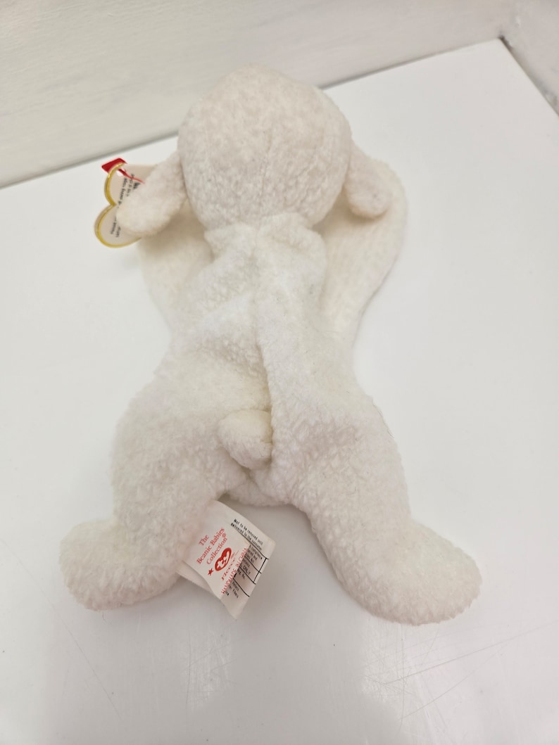 May include: A white plush lamb toy from The Beanie Babies Collection. The lamb has a soft, textured exterior, floppy ears, and a small tail. A tag is attached to the lamb's ear. The toy is lying on a white surface.